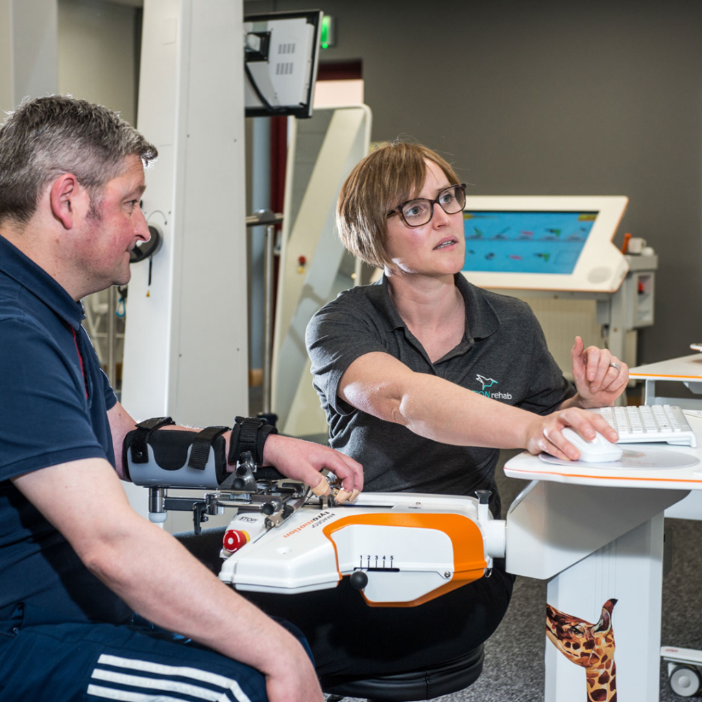 A woman sits with a man using a device to help the man with neuro rehabilitation, the device is attached to the mans hand, the woman looks at a screen