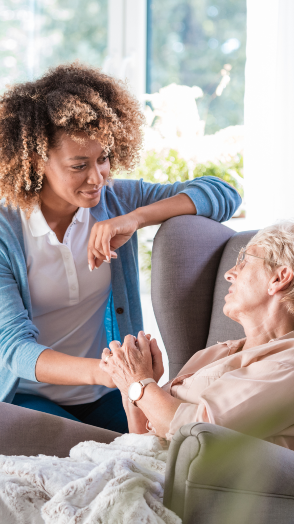 An image of a young lady holding the hand of an older lady that sits in a chair, insinuatin care and a friendly relationship